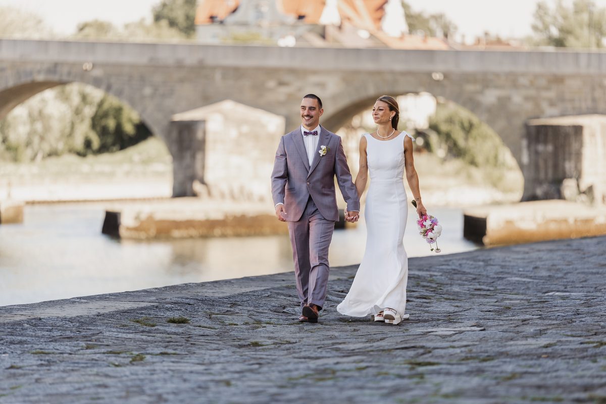 Brautpaar geht Hand in Hand am Donauufer in Regensburg, im Hintergrund die Steinerne Brücke