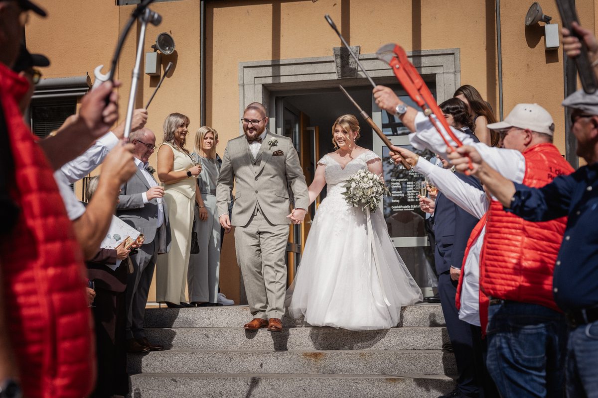 Christina und Dominik kommen Hand in Hand die Treppe vom Standesamt Maxhütte-Haidhof herunter, Gäste halten Rohrzangen und Schraubenschlüssel als Spalier hoch