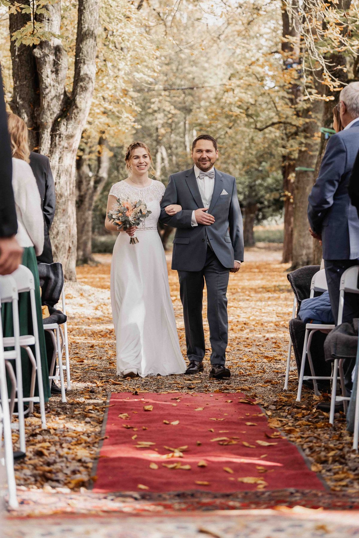 Linda und Oliver beim gemeinsamen Einzug zur freien Trauung auf rotem Teppich durch die herbstliche Allee im Prüfeninger Schlossgarten